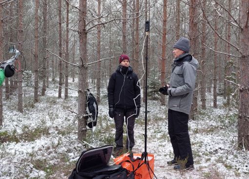 Kuvassa on kolme henkilöä talvisessa metsässä tuottamassa live-lähetystä Helsingin yliopiston opiskelijoille